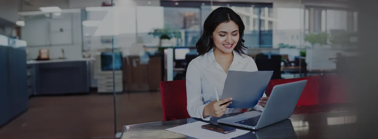 Person sitting at a desk reviewing documents while working on a laptop in an office setting banner from ICICI bank.