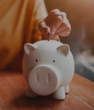Person placing a coin into a white piggy bank on a table to represent saving money | ICICI Bank